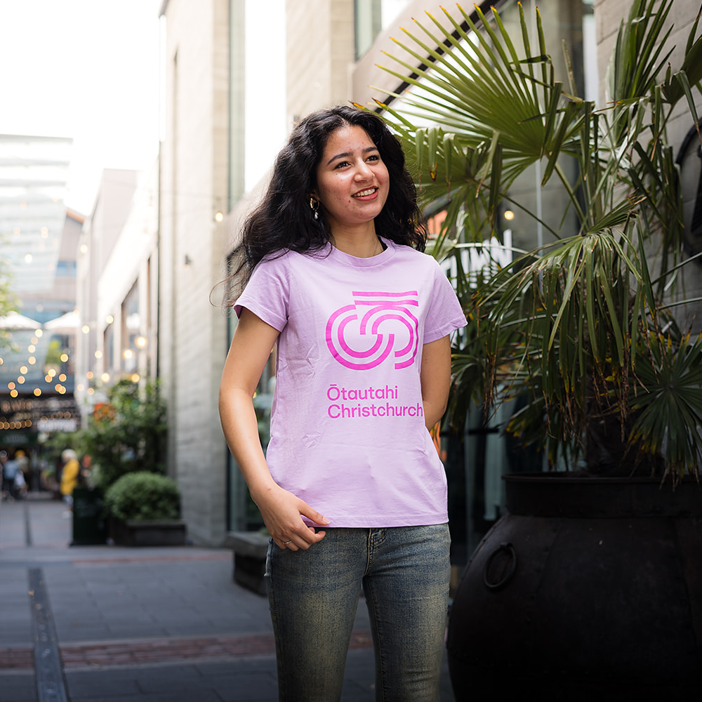 Person wearing a pink t-shirt with a logo and text, standing outdoors.