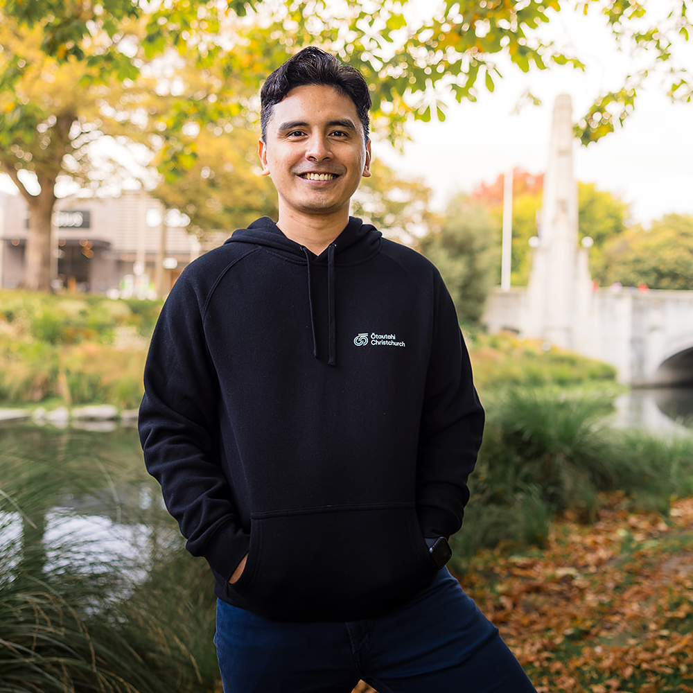 Man wearing a black hoodie with a logo standing outdoors near a pond and trees.