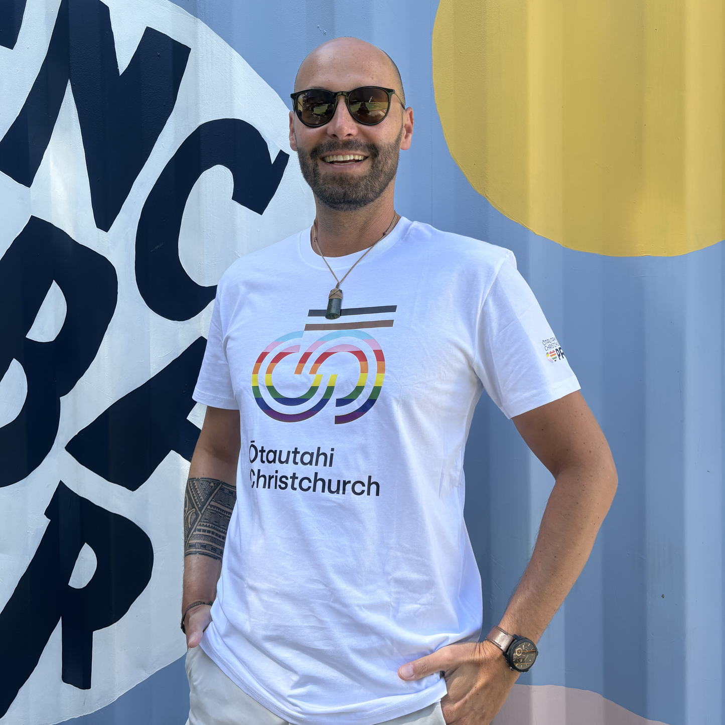 Man wearing a white t-shirt with rainbow design and text, standing in front of a blue wall with large letters.
