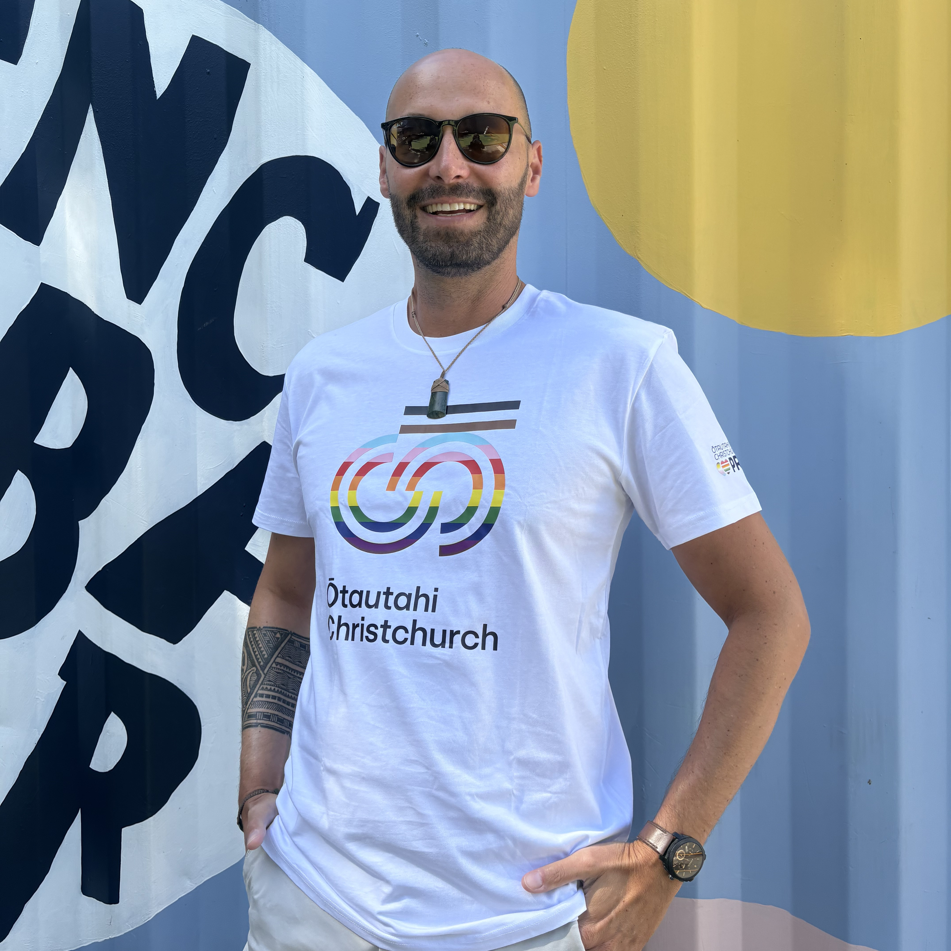 Man wearing a white t-shirt with rainbow design and text, standing in front of a blue wall with large letters.