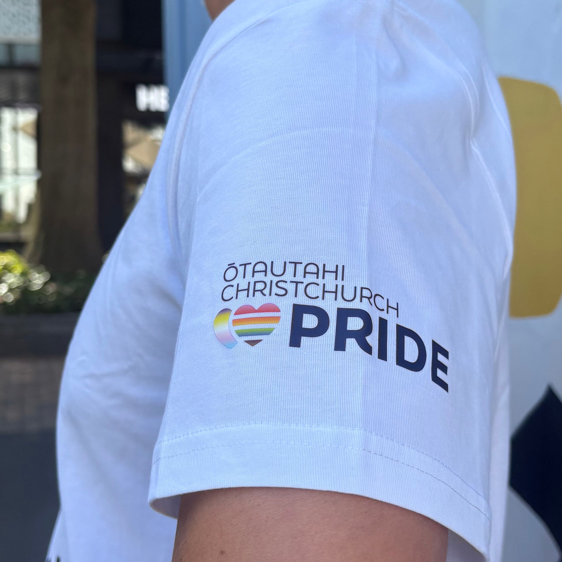 White t-shirt with 'ŌTAUTahi Christchurch Pride' text and rainbow hearts on a blurred background